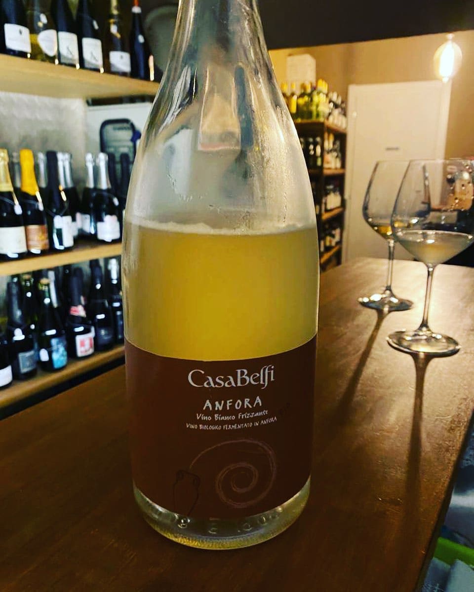 Bottiglia di CasaBelfi Anfora, vino bianco frizzante, sul bancone, con scaffali di bottiglie e due calici sullo sfondo