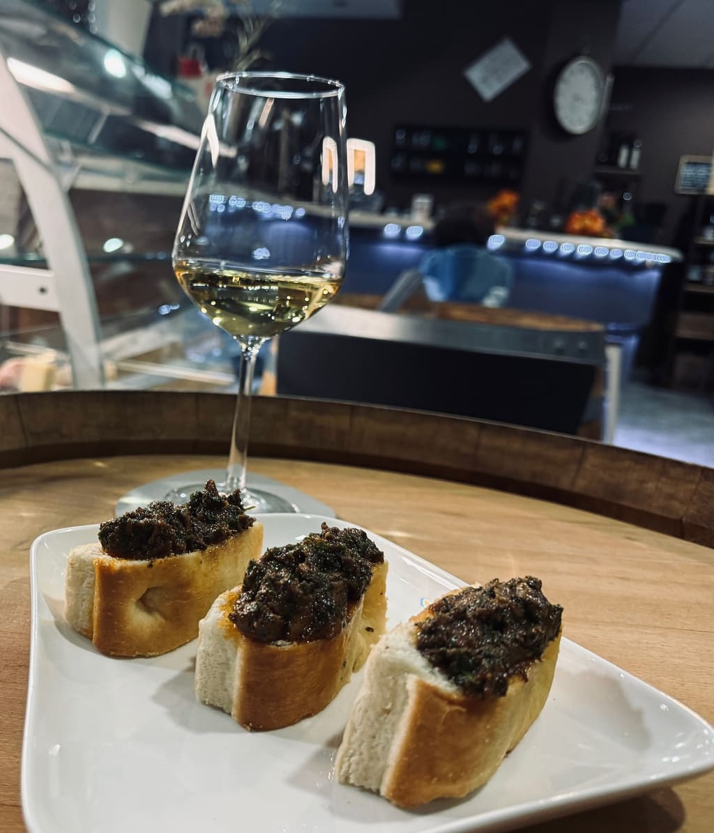 Tre crostini e un calice di vino bianco su un tavolo a botte, il bancone del locale sullo sfondo