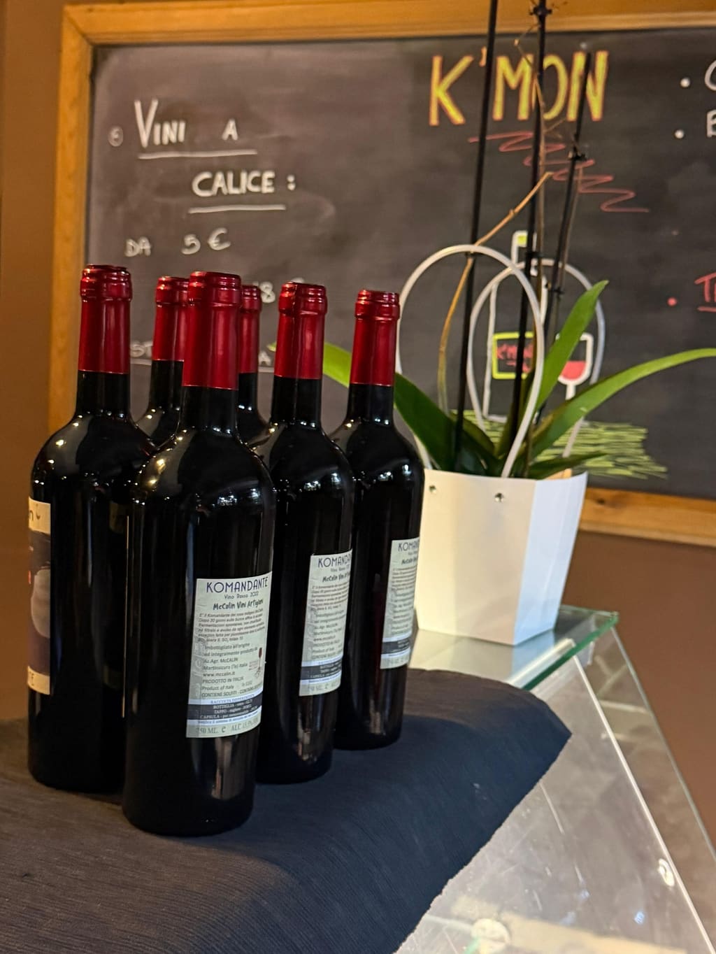 Bottiglie di Komandante, rosso di McCalin Vini Artigiani, davanti alla lavagna con i vini a calice da 5 €