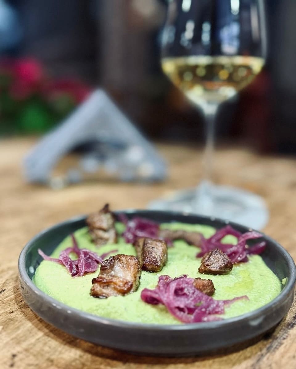 Crema verde di piselli con cubetti di carne rosolata e cipolla rossa, accanto a un calice di bianco